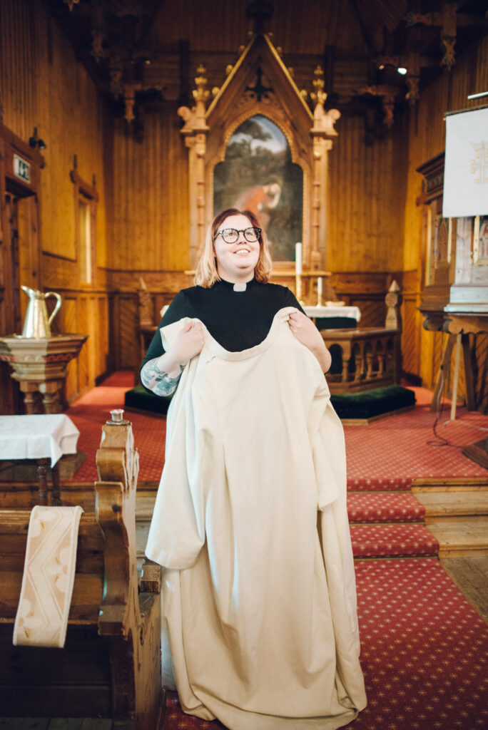 Prest Silje Mathea står i kirkegangen og ser opp mot organistetn. Hun holder prestekjolen i hendene