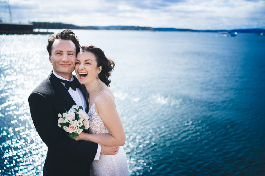 Brudepar fotografert på Aker Brygge med Oslo-fjorden i bakgrunnen. De holder rundt hverandre og smiler naturlig.